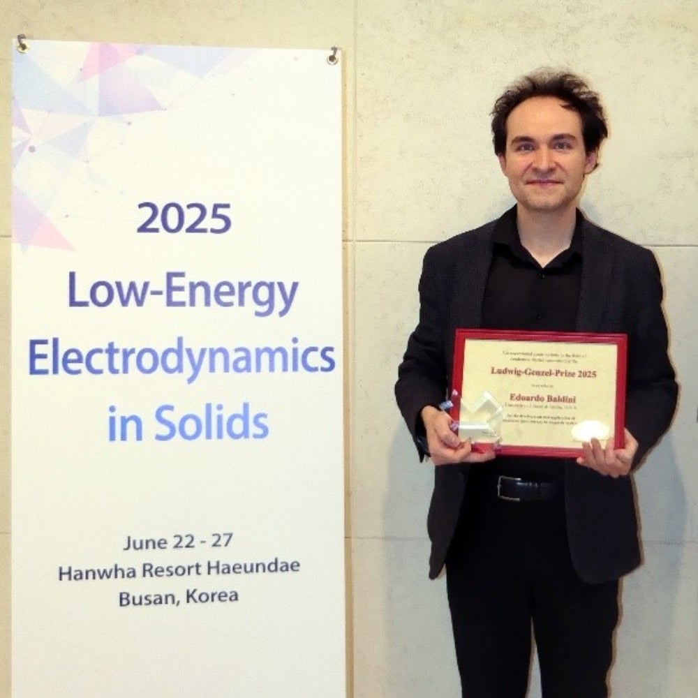 A man clad in black holds a certificate, standing next to an upright banner that reads: 2025 Low-Energy Electrodynamics in Solids, June 22-27, Hanwha Resort Haeundae, Busan, Korea