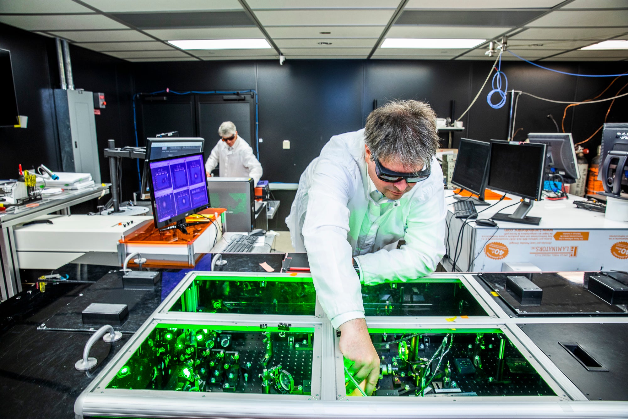 A scientist wearing protective goggles leans over a laser in a lab filled with cutting-edge equipment.